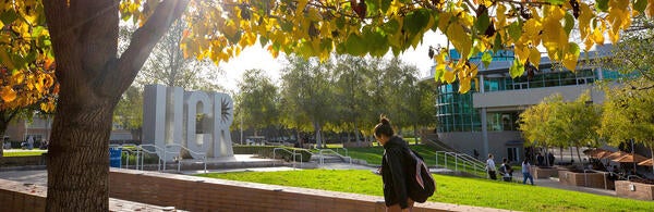 Student walking by UCR sign under trees in the fall